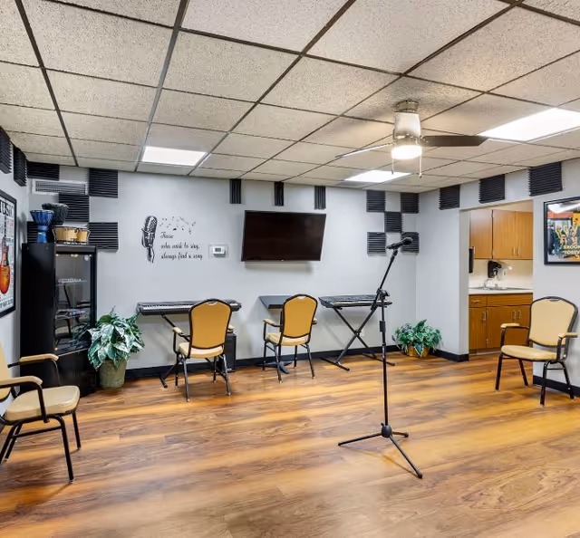 A music room with wooden flooring and a ceiling fan. The room has three keyboards on stands against the wall, each with a chair in front. A microphone stand is positioned in the center of the room. Acoustic foam panels are mounted on the walls around a flat-screen TV. There are plants in pots on the floor and a small kitchenette visible through an open doorway. The wall has a quote that reads, 'Those who wish to sing, always find a song.'