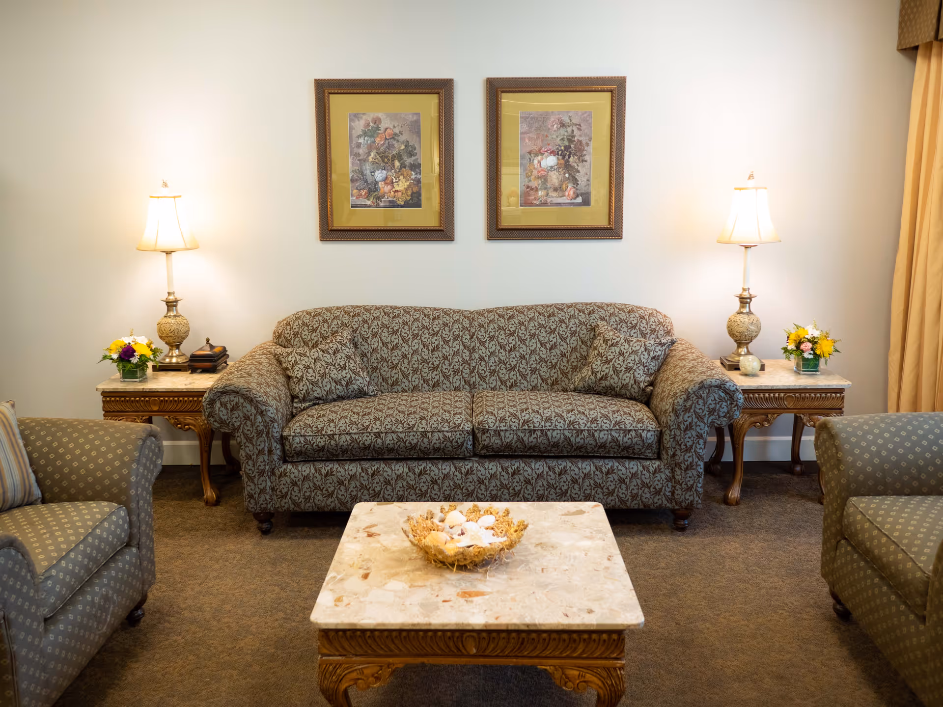 A traditional living room with a patterned sofa, two armchairs, side tables with lamps and flowers, framed artwork above, and a marble-topped coffee table.