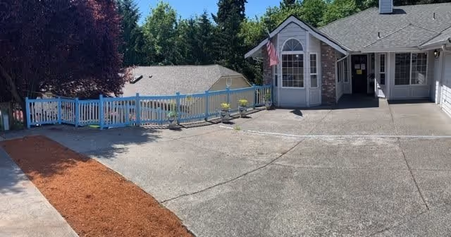 Exterior view of a senior care facility showing a paved driveway leading to the entrance of a single-story building with a gray roof and white siding. There is a white fence with flower pots along it, an American flag mounted near the entrance, and trees in the background under a clear blue sky.
