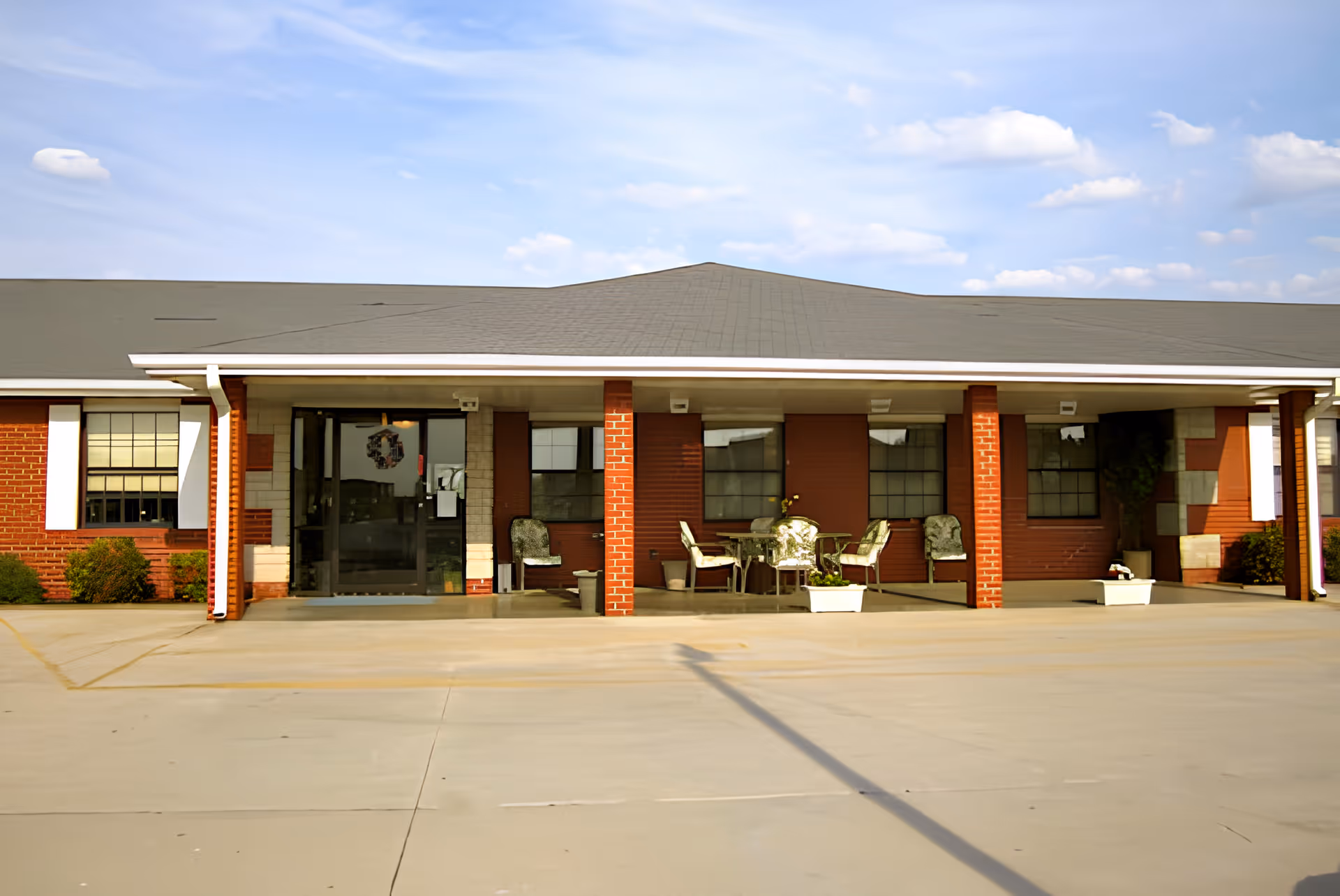 Single-story brick building entrance with a covered porch, outdoor seating and potted plants under a blue sky.