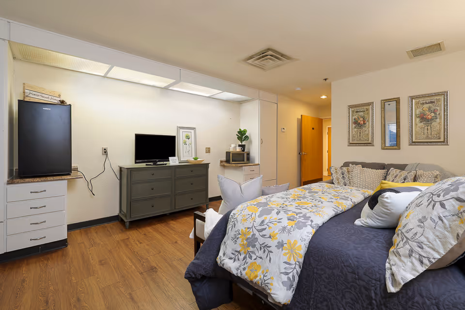 A cozy bedroom in an assisted living facility featuring a bed with floral and patterned bedding and multiple pillows. The room has wooden flooring, a gray dresser with a small TV and framed artwork on top, a mini refrigerator, a microwave, and a small plant. The walls are decorated with two framed floral pictures and a mirror. A doorway leads to another part of the facility.