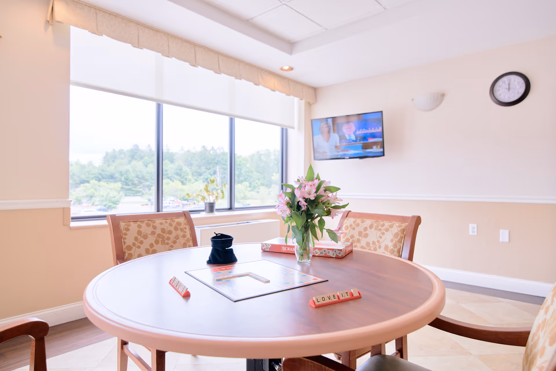 A bright room with a round wooden table set for a game of Scrabble, featuring a vase of pink flowers in the center. Four cushioned chairs with floral upholstery surround the table. Large windows with white blinds let in natural light and offer a view of green trees outside. A wall-mounted TV and a clock are visible on the beige and white walls.