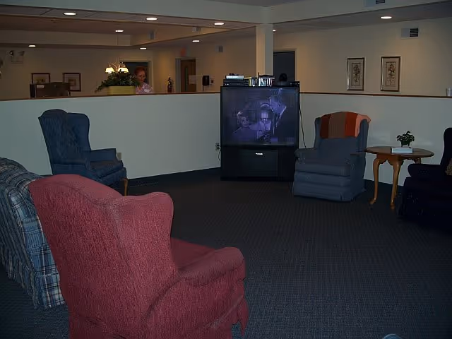 Communal living room with several upholstered armchairs arranged around a television and a low partition wall.