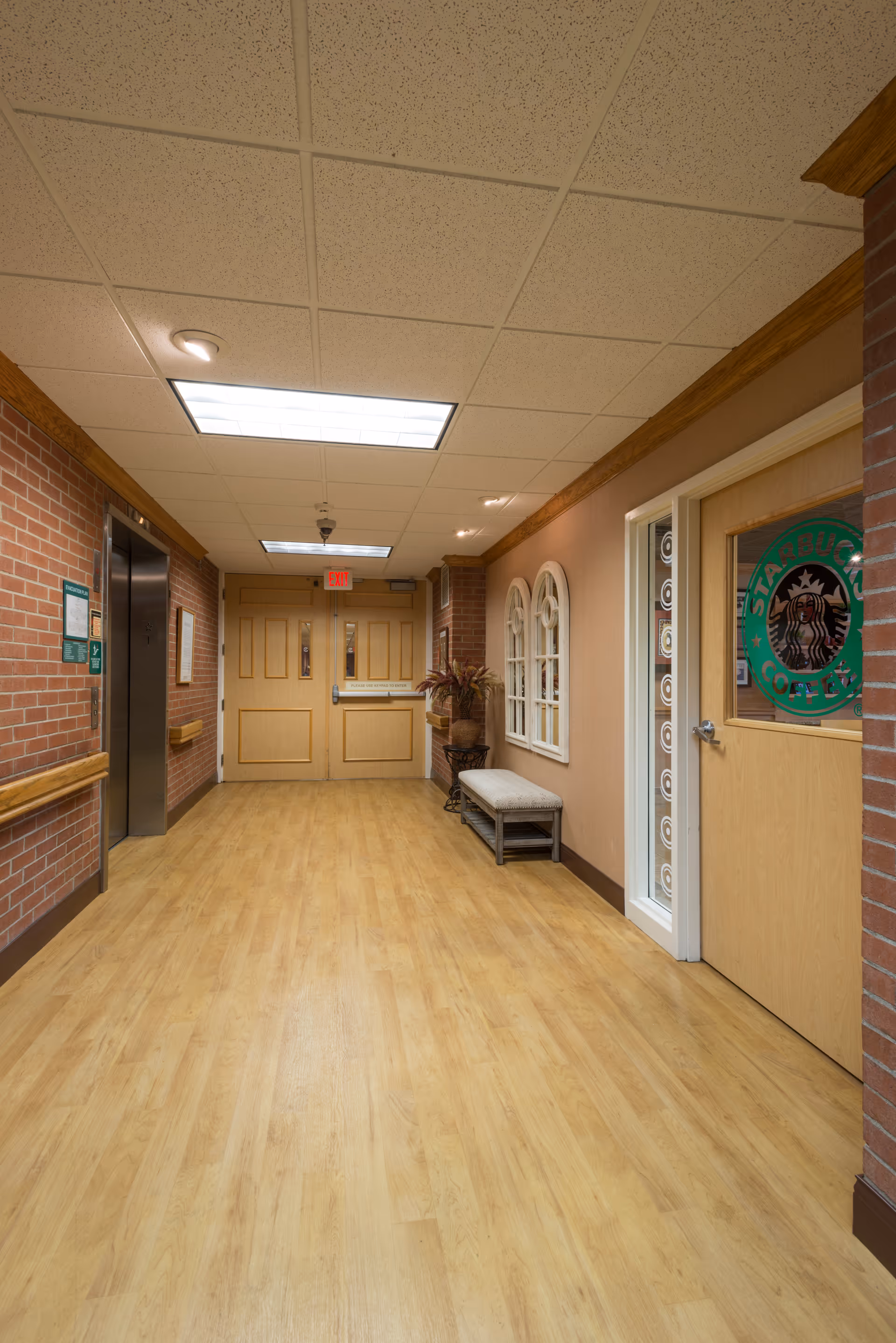 Well-lit interior corridor with wood floors, an elevator on the left, a bench and decorative wall windows on the right, and double doors at the far end.