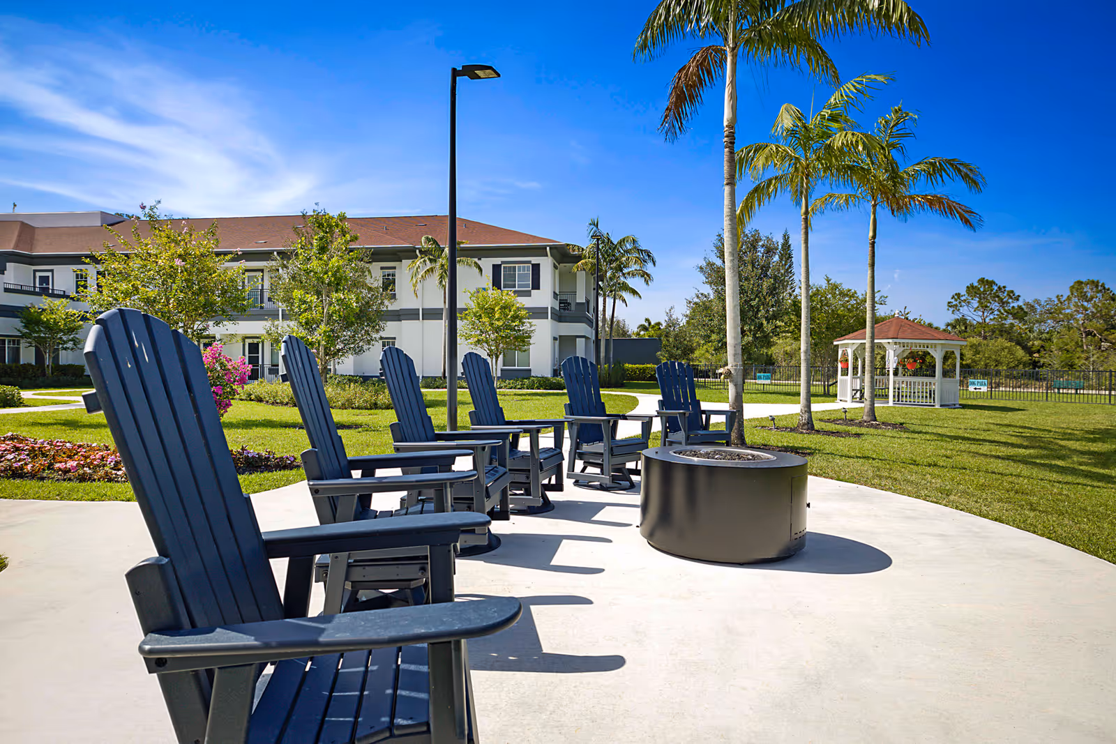 Outdoor seating area at Alamar Senior Living with several dark blue Adirondack chairs arranged around a circular fire pit. The area is surrounded by green grass, palm trees, and a white gazebo in the background under a clear blue sky.