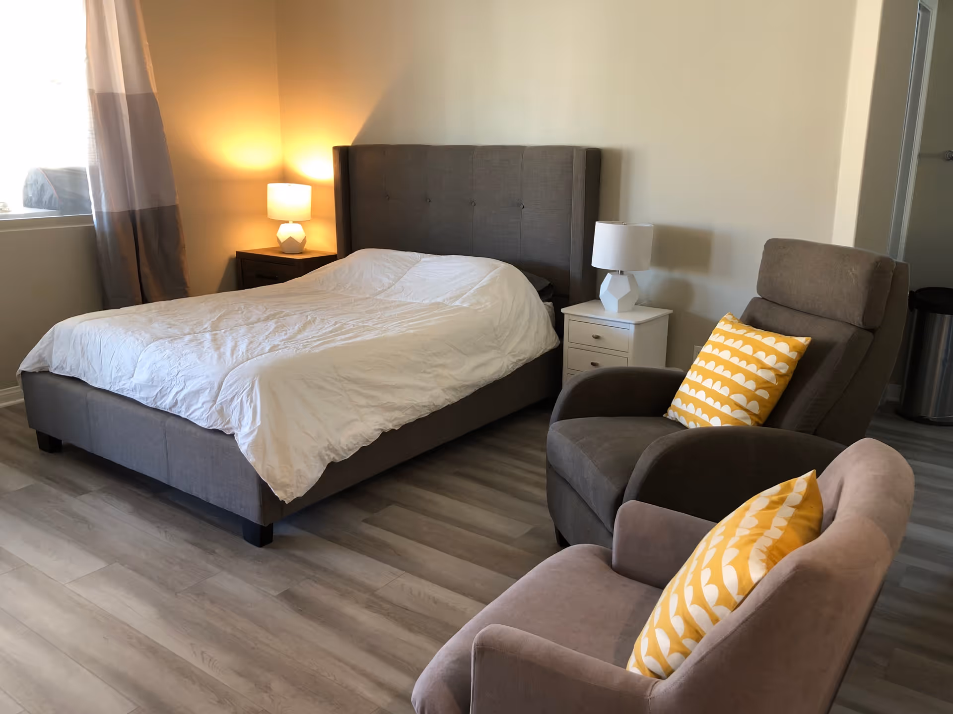 A bedroom with a gray upholstered bed with white bedding, two nightstands each with a white lamp, and two armchairs with yellow patterned pillows. The room has light wood flooring and a window with curtains letting in natural light.