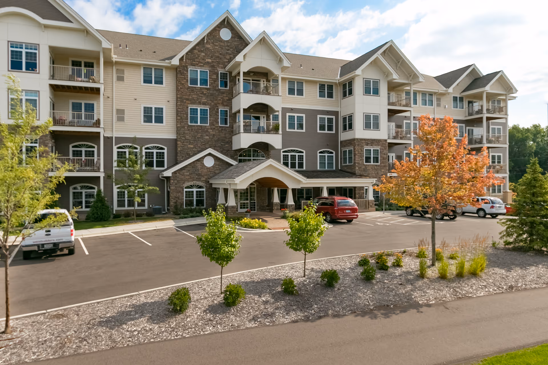 Exterior view of a multi-story senior living facility building with balconies, a covered entrance, parked cars, and landscaped greenery including small trees and shrubs.