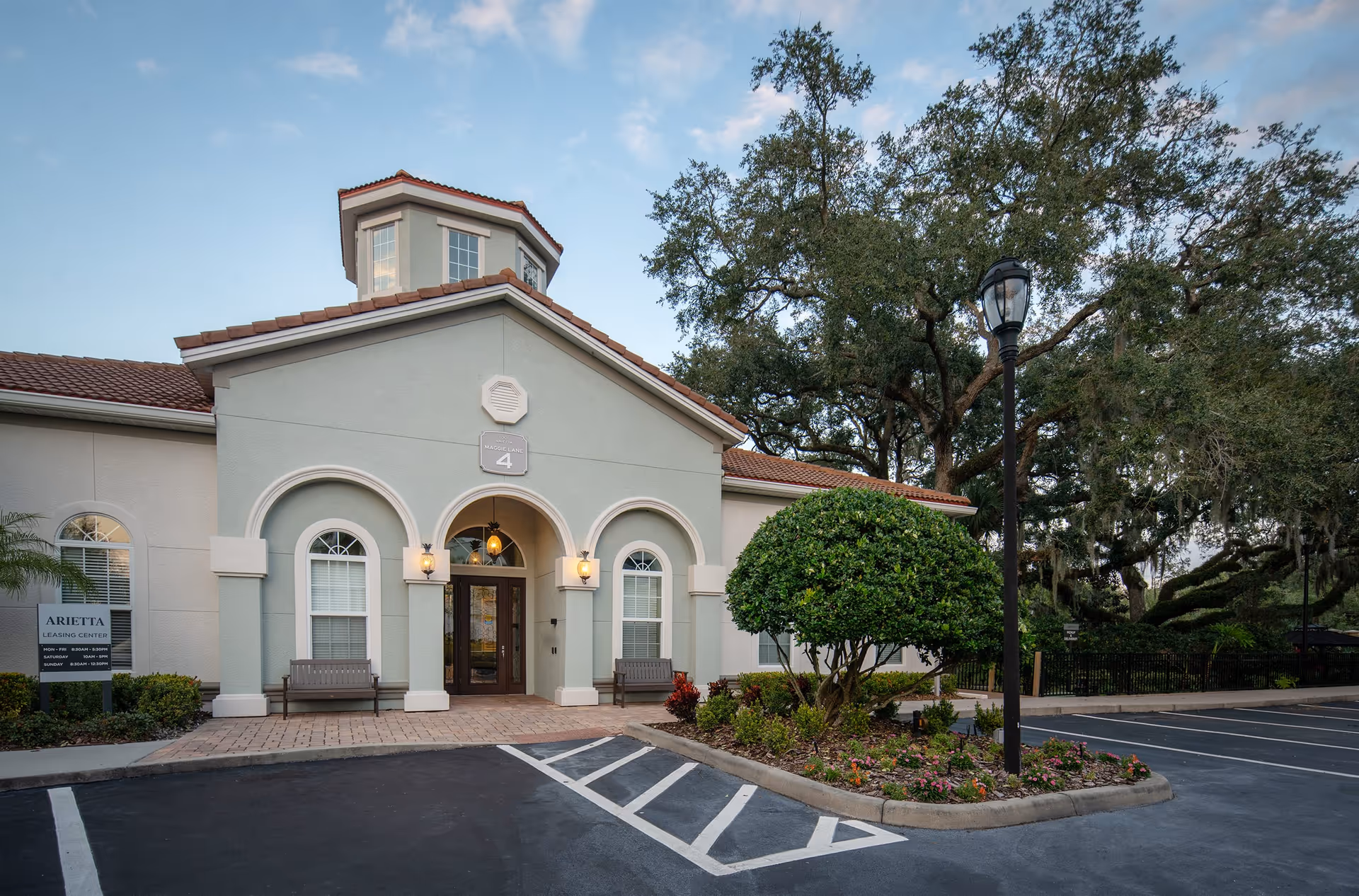 Exterior view of the Arietta leasing center building with a light green facade, arched windows and entrance, two benches outside, a landscaped area with a trimmed bush and flowers, a street lamp, and a parking lot in front under a partly cloudy sky.
