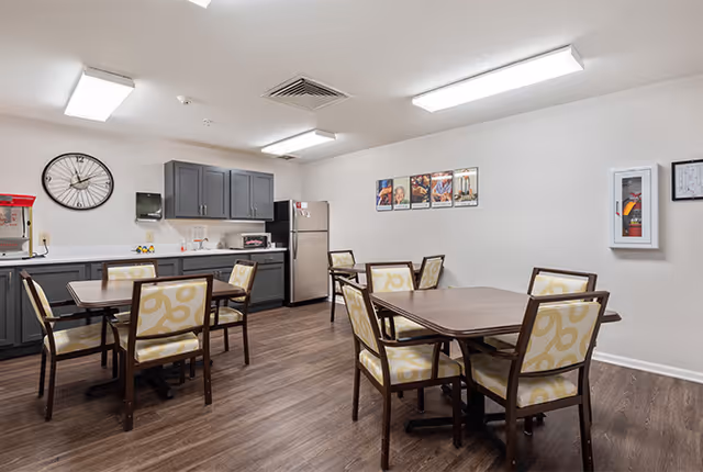 A dining area in an assisted living facility featuring three wooden tables each surrounded by four cushioned chairs with patterned upholstery. The room has wood flooring, white walls, and a ceiling with fluorescent lighting. Along one wall, there is a kitchenette with gray cabinets, a stainless steel refrigerator, a microwave, a popcorn machine, and a large wall clock. A fire extinguisher is mounted on the opposite wall, and framed pictures hang above the kitchenette.