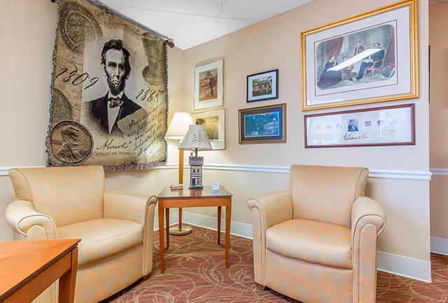 A cozy sitting area with two beige armchairs facing each other, separated by a wooden side table with a decorative lamp. The walls are adorned with framed pictures and a large tapestry featuring an image of Abraham Lincoln and the years 1809 and 1865. The carpet has a patterned design in warm tones.