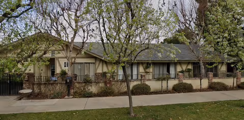 Exterior view of a single-story residential building with beige walls and a brown roof, partially obscured by leafless trees and shrubs. A sidewalk runs in front of the building, which is enclosed by a low fence.