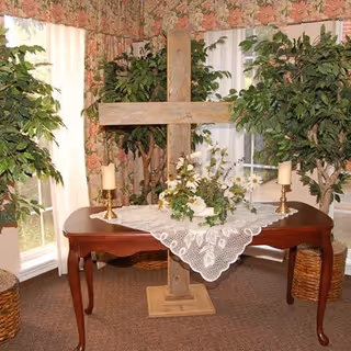 A wooden cross displayed on a dark wooden table covered with a white lace tablecloth, flanked by two gold candle holders with white candles. The table is set in a corner with floral wallpaper, two large potted green plants, and windows with white curtains.