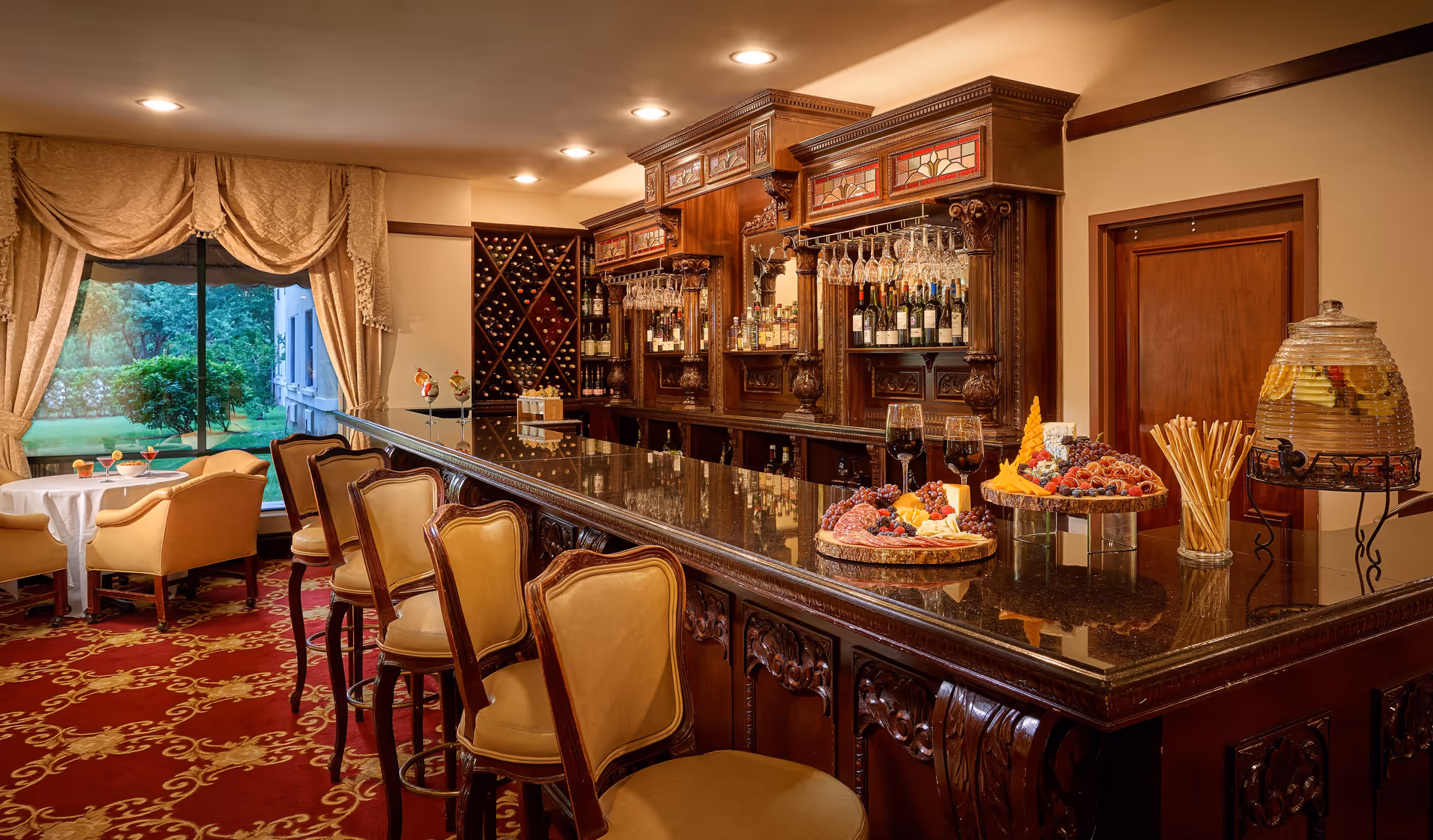 An elegant indoor bar area with a polished dark granite countertop and ornately carved wooden cabinetry. Four cushioned bar stools are lined up along the counter. Behind the bar, there are shelves stocked with wine bottles and hanging wine glasses. On the counter, there are platters of assorted fruits and snacks, two glasses of red wine, and a large glass dispenser filled with a fruit-infused beverage. To the left, there is a seating area with two beige armchairs and a small round table covered with a white tablecloth, positioned near a large window with beige drapes that overlook a green outdoor garden.