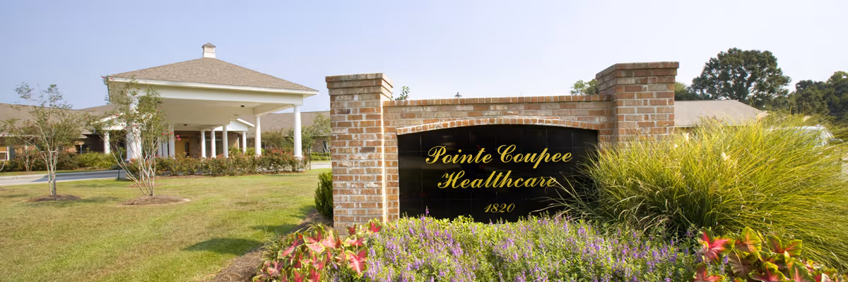 Brick entrance sign reading 'Pointe Coupee Healthcare' with landscaped flowers and the facility's covered entrance in the background.
