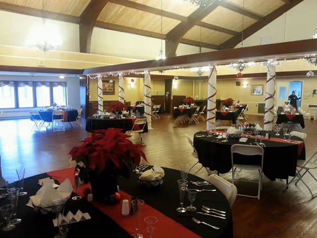 A spacious dining room decorated for a festive occasion with round tables covered in black tablecloths and red runners. Each table is set with glassware, silverware, napkins, and a centerpiece featuring red poinsettias. The room has wooden floors, high ceilings with exposed beams, and white columns wrapped with red and white garlands and string lights. Large windows allow natural light to enter the room.