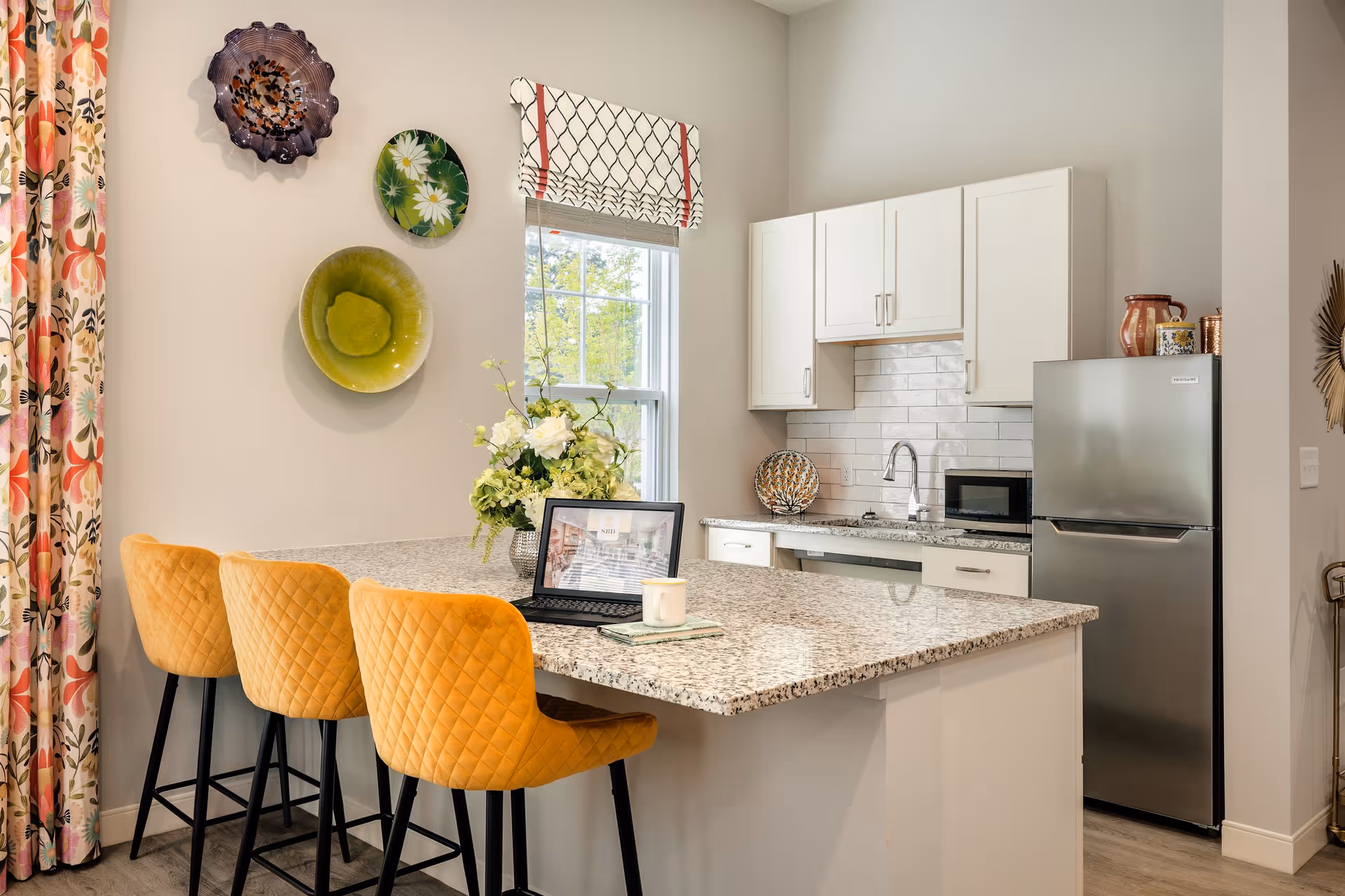 Bright kitchen with a granite island, three yellow quilted bar stools, white cabinets, a stainless steel refrigerator, and decorative plates on the wall.