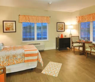 A cozy bedroom with a bed covered in patterned orange and white bedding, two windows with orange valances, a small black dresser with a lamp and decorative items, and a round table with two chairs near the windows. The room has wooden flooring and light-colored walls.