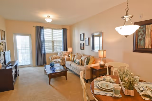A cozy living room and dining area with beige walls and carpet. The living room features a beige sofa with multiple cushions, a patterned armchair, a wooden coffee table with decorative items, and a TV on a stand. The dining area has a round wooden table set with plates, cups, and napkins, along with a hanging light fixture above. There are two table lamps on side tables beside the sofa, a window with blue curtains, and a door with glass panes letting in natural light.
