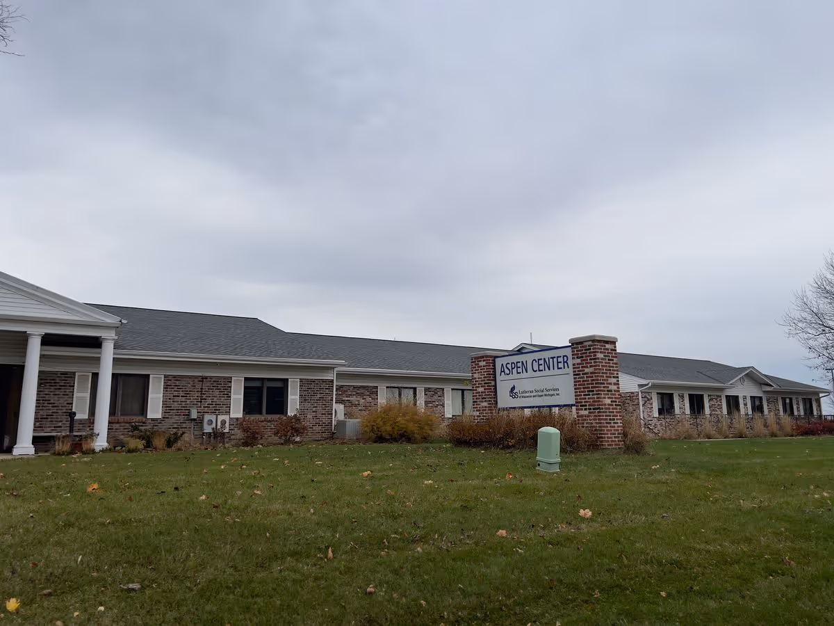 Low single-story brick building with a lawn and a sign reading "Aspen Center" under an overcast sky.
