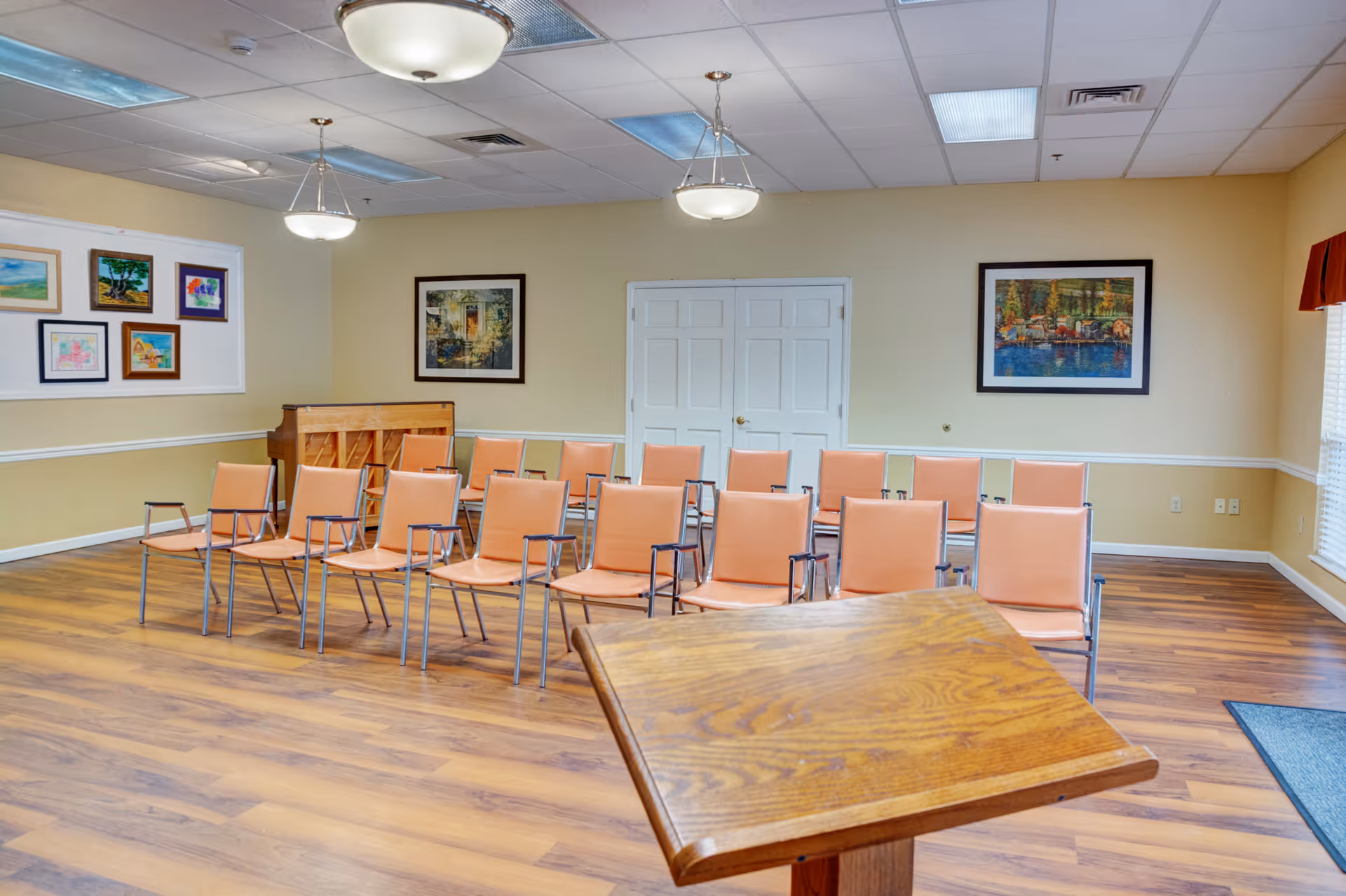 A room with rows of peach-colored chairs arranged facing a wooden podium. The room has light yellow walls adorned with framed artwork and a piano in the corner. The floor is wooden, and there are ceiling lights and a window with blinds and a red valance on the right side.