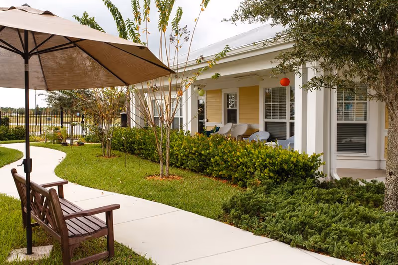 Outdoor patio area at The Brennity at Tradition Senior Living featuring a curved concrete walkway, wooden bench with a large umbrella, green grass, small trees, shrubs, and a building porch with chairs and windows.
