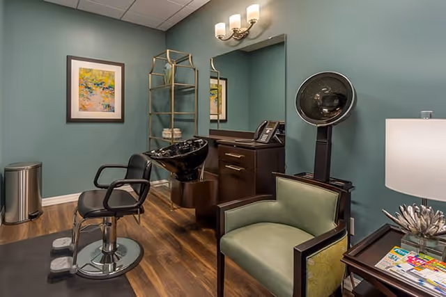 Interior view of a salon area with a black salon chair in front of a black hair washing basin, a green armchair, a wooden cabinet with a phone, a wall mirror, a metal shelving unit, a framed colorful painting on a teal wall, and a floor lamp next to a small table with magazines.