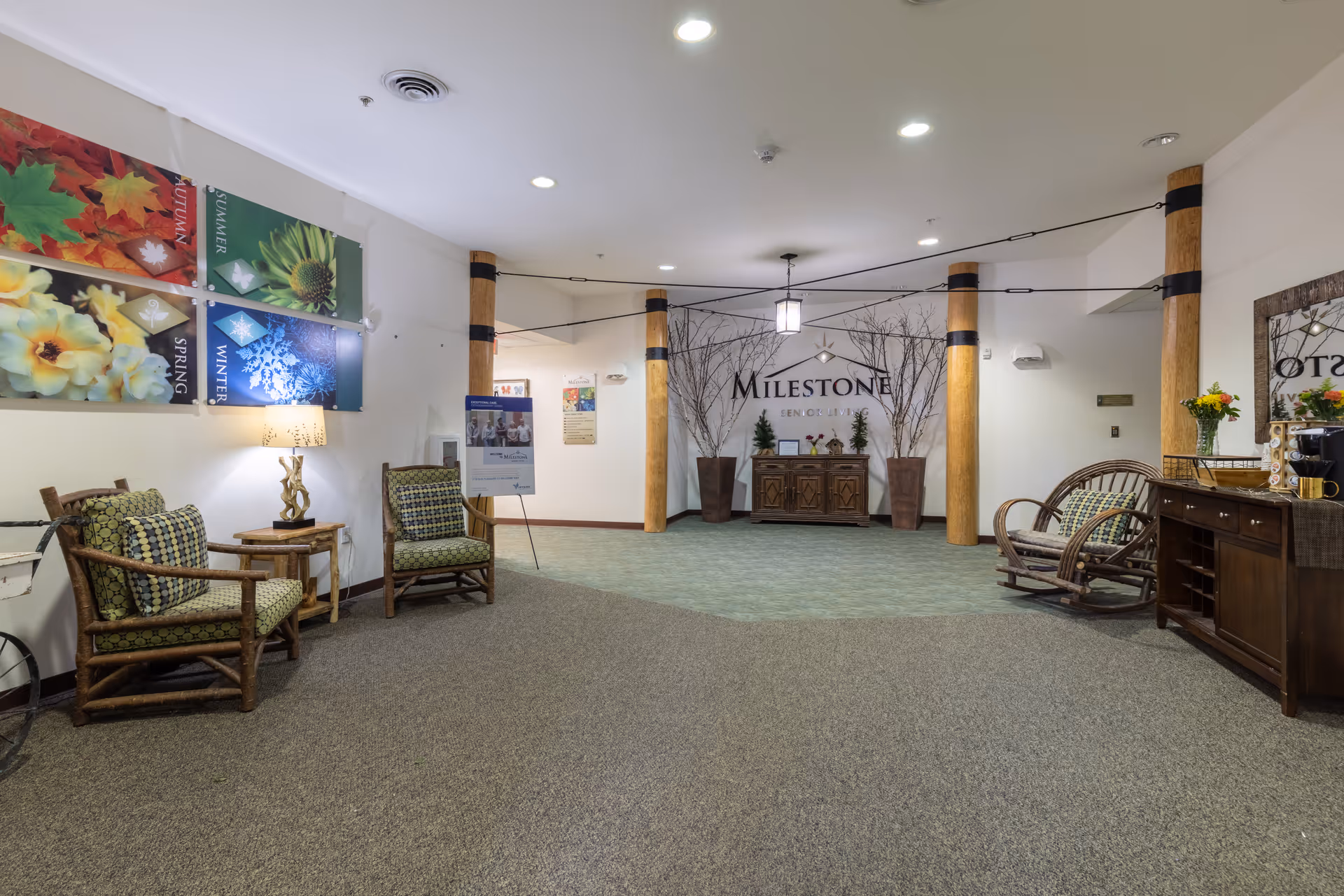 Interior view of a senior living facility lobby area with wooden chairs featuring green patterned cushions, a wooden rocking chair, a side table with a lamp, and a wooden cabinet with flowers and a coffee maker. The back wall displays the Milestone Senior Living logo with decorative tall vases and branches. Colorful seasonal flower artwork is mounted on the left wall.