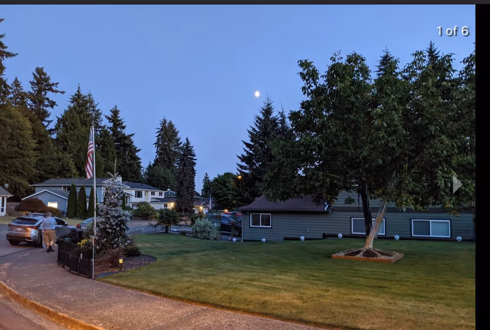 Outdoor view of a residential area at dusk with a lawn, trees, and houses. A person is walking near a parked car on the street. The sky is clear with the moon visible.