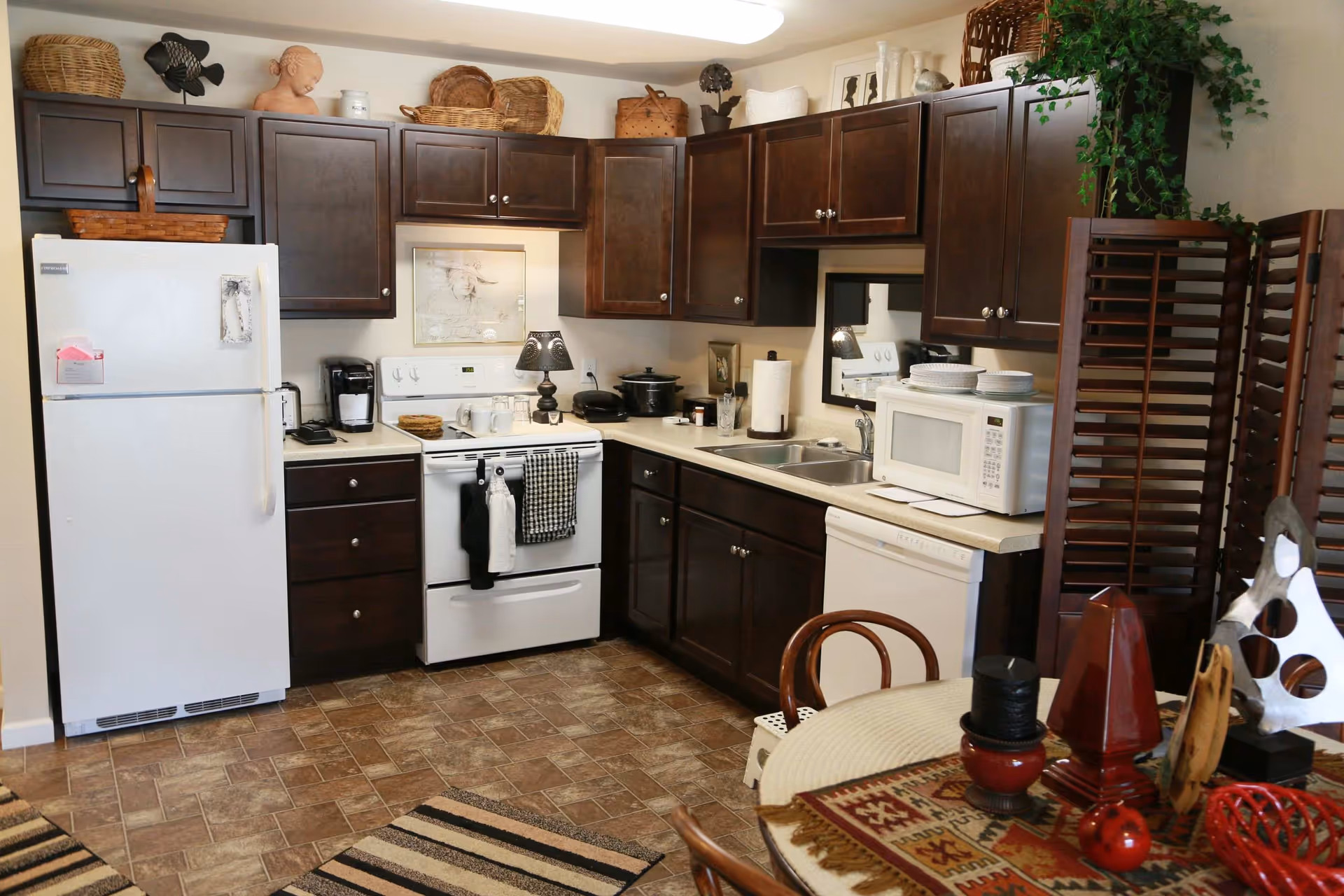 A kitchen with dark wooden cabinets, a white refrigerator, white stove, microwave, and dishwasher. The countertops have various kitchen items including a coffee maker, paper towel holder, and dishes. There is a small round dining table with decorative items and chairs in the foreground. The floor has a patterned tile and there are woven baskets and decorative pieces on top of the cabinets.