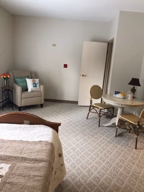 A cozy room with a bed partially visible in the foreground, a beige armchair with a decorative pillow and a small side table with flowers in the corner, a round table with two patterned chairs, and a lamp on the table. The walls are light-colored and the carpet has a diamond pattern. A door is open in the background.