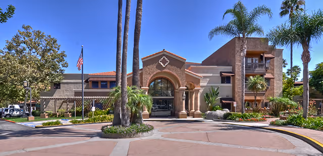 Exterior front view of a senior living facility named Ivy Park of Wellington, featuring a large entrance with an archway, palm trees, landscaped greenery, and a clear blue sky.