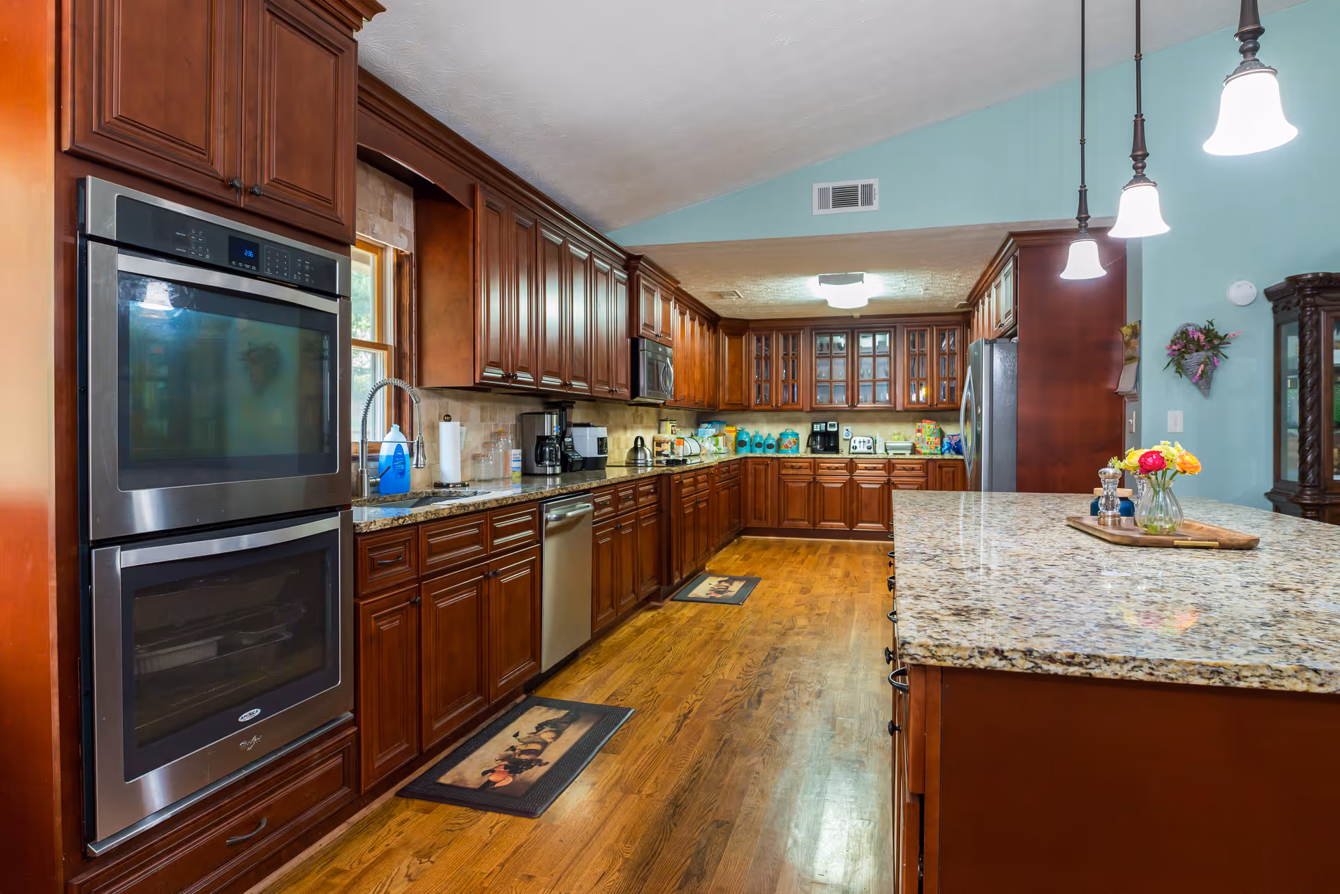 Spacious kitchen with wooden cabinets, granite countertops, double wall ovens, stainless steel dishwasher and refrigerator, hardwood floors, and pendant lights hanging over a large kitchen island with a flower vase on top.