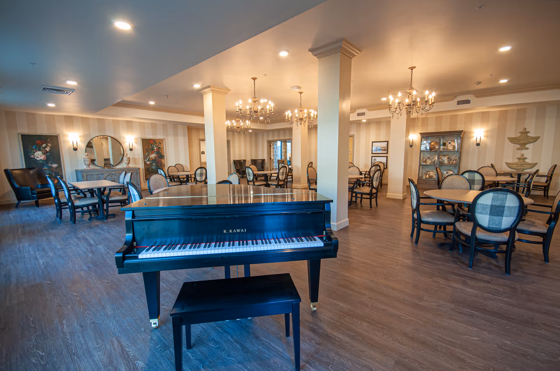 A spacious senior living community room with a black K. Kawai grand piano and matching bench in the foreground. The room features multiple round tables with cushioned chairs, elegant chandeliers hanging from the ceiling, and decorative wall art including paintings and a mirror. The flooring is wood-style, and the walls have a striped pattern with soft lighting fixtures.