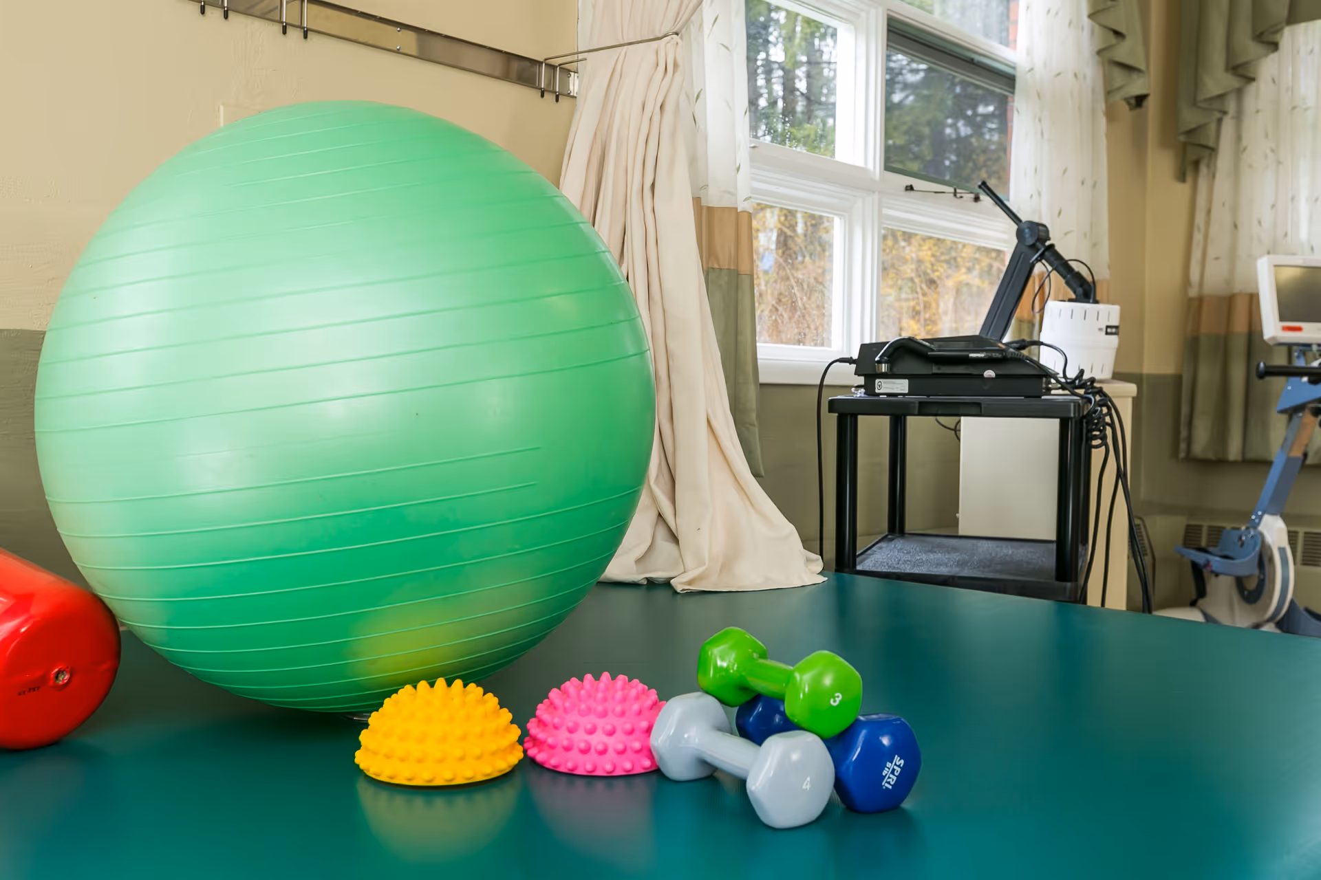 Rehabilitation room with a large green exercise ball, small dumbbells and textured balance pads on a table beside windows and therapy equipment.