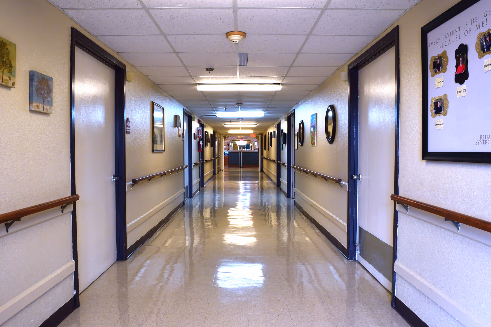 A long, clean hallway in a senior living or nursing facility with handrails on both sides, several closed doors, framed pictures and mirrors on the walls, and bright overhead lighting.