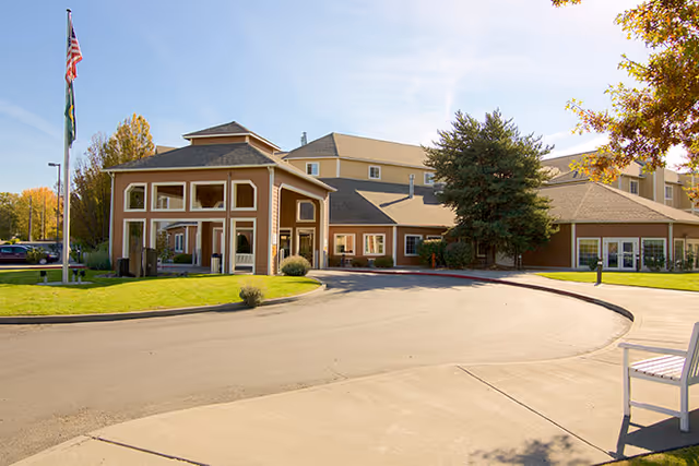 Exterior view of Brookdale Richland senior living facility on a sunny day, showing the main entrance with a covered drop-off area, well-maintained lawn, trees, and an American flag on a flagpole.