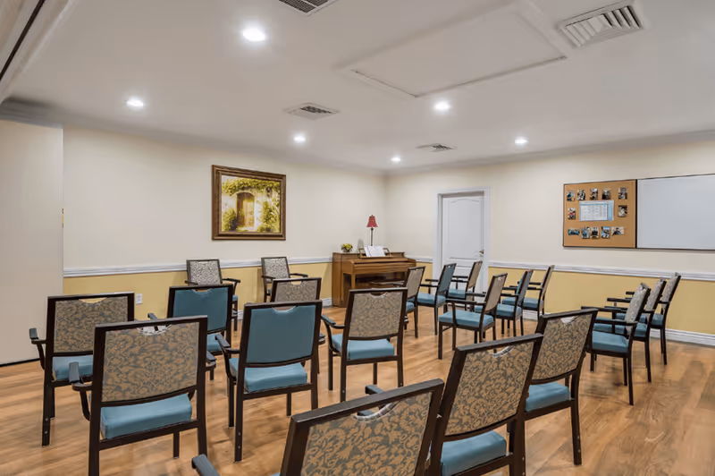 A room with multiple chairs arranged in rows facing a piano against the wall. The room has wooden flooring, beige walls with a white and yellow trim, a framed painting, a bulletin board with photos and papers, and a whiteboard. The ceiling has recessed lighting and air vents.