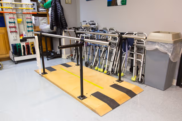 A rehabilitation or physical therapy area with parallel bars mounted on a wooden platform for walking exercises. Behind the bars, there are several walkers lined up against the wall, a trash bin with a plastic liner, and a rack holding various exercise bands and equipment. The floor is light blue and the walls are light-colored.