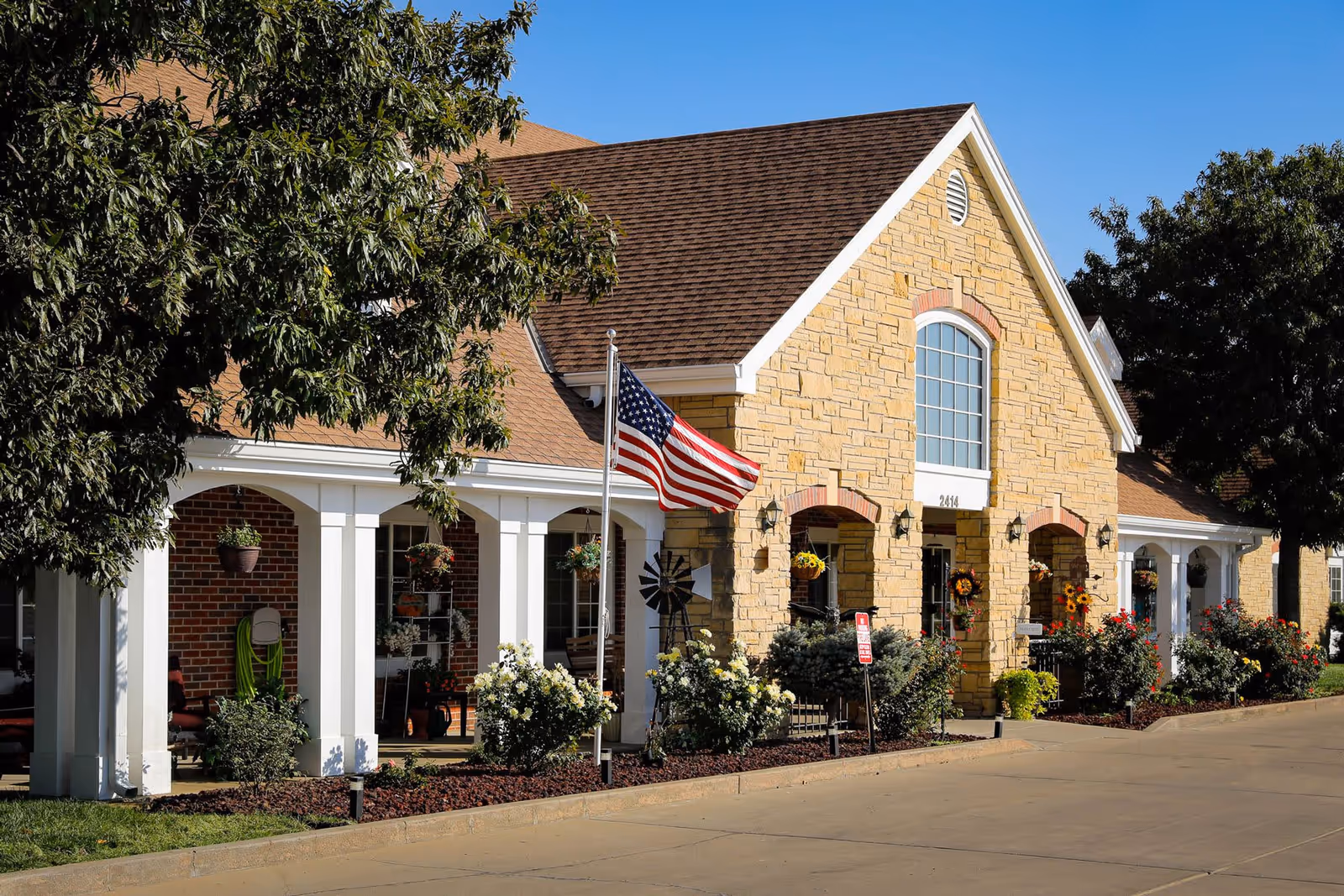 Exterior view of Homestead Assisted Living of Garden City building with stone and brick facade, white columns, an American flag on a flagpole, and landscaped flower beds with bushes and hanging plants under a clear blue sky.