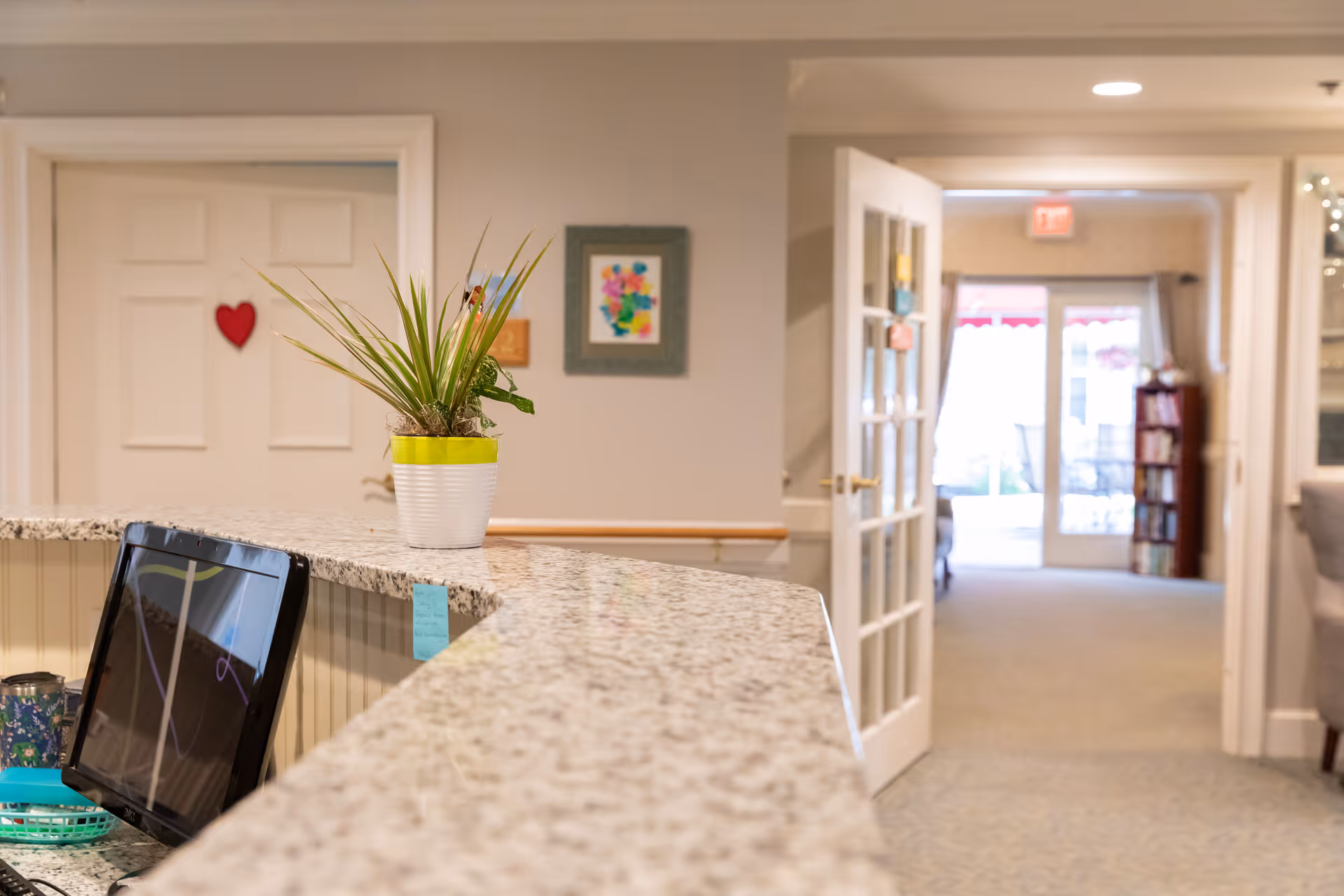 A reception counter with a potted plant on a granite countertop overlooking an open hallway and glass doors inside a senior living facility.