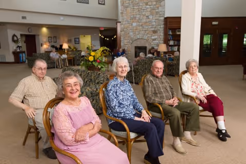 Five elderly individuals sitting on wooden chairs arranged in a semi-circle in a spacious, well-lit common area with a stone fireplace, bookshelves, and floral arrangements in the background.