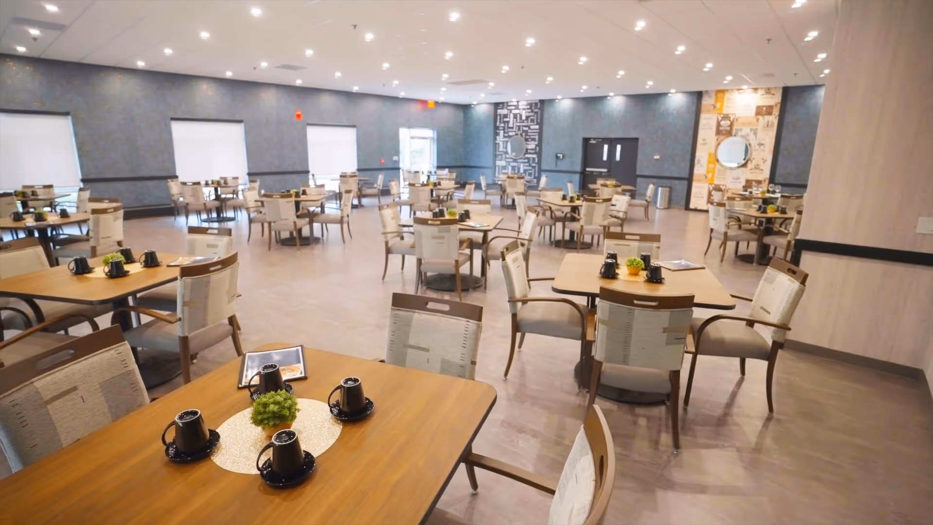 A spacious dining room with multiple wooden tables and chairs arranged neatly. Each table has black coffee mugs and small green plants as centerpieces. The room has large windows with white blinds, recessed ceiling lights, and a modern decor with blue and beige walls.