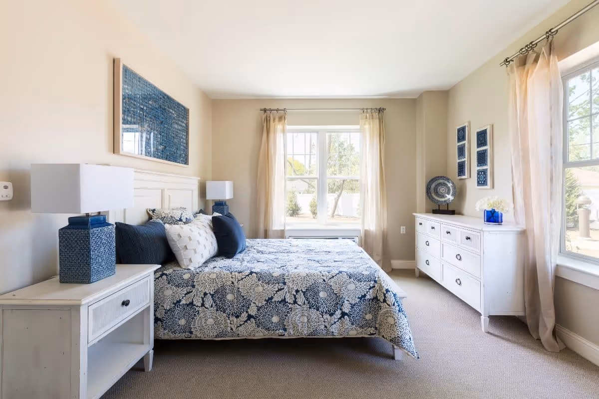 A bright and airy bedroom with a large bed covered in a blue and white floral patterned comforter and multiple pillows. The room features beige walls, two large windows with sheer curtains allowing natural light to fill the space, a white nightstand with a blue lamp, and a white dresser adorned with decorative items including a blue vase and framed artwork on the walls.