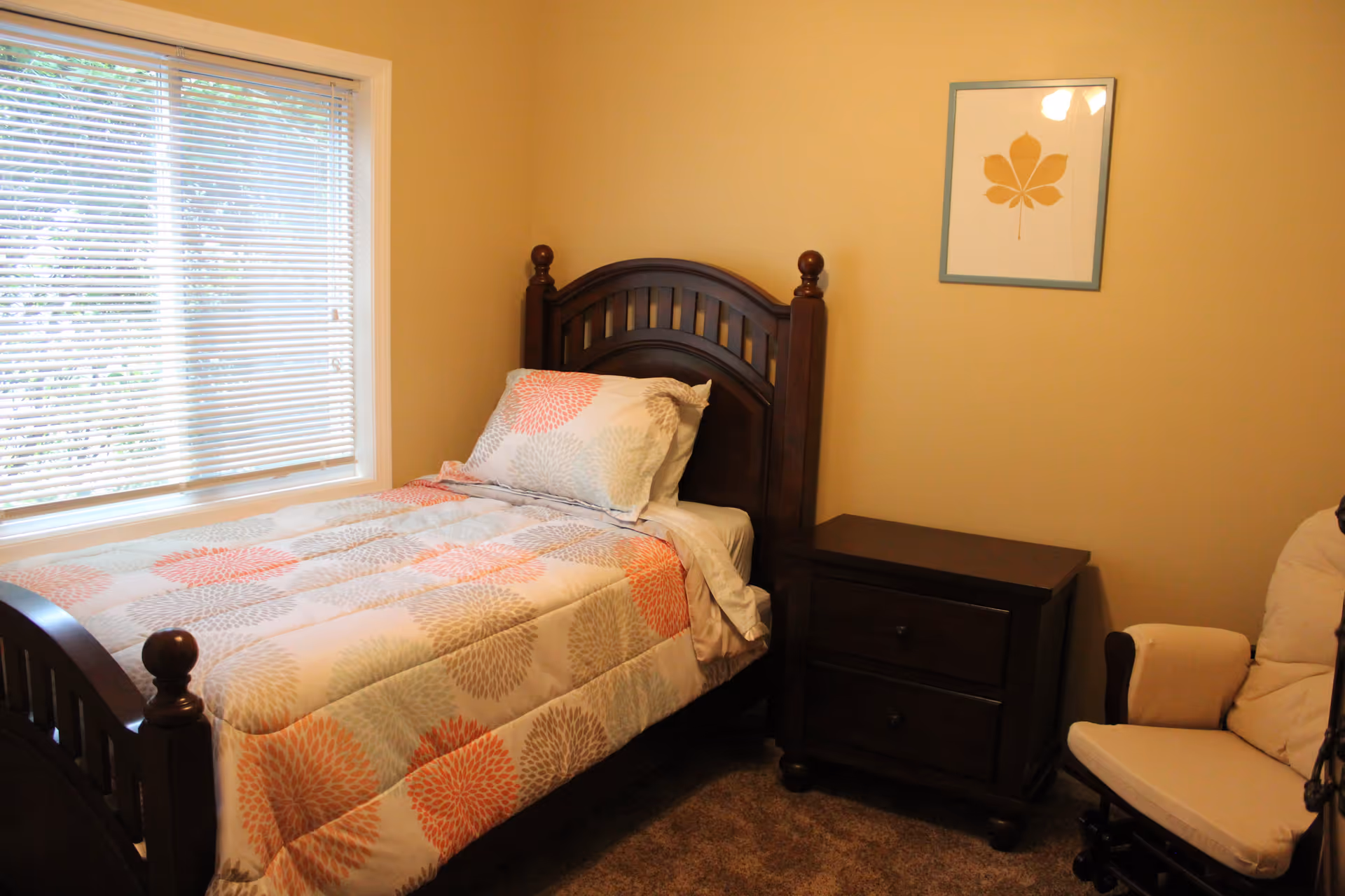 A small bedroom with a single wooden bed featuring a floral patterned comforter and pillow. Next to the bed is a dark wooden nightstand with two drawers. A cushioned beige armchair is partially visible on the right. A framed picture of a leaf hangs on the beige wall above the nightstand. A window with white blinds is on the left side of the room.