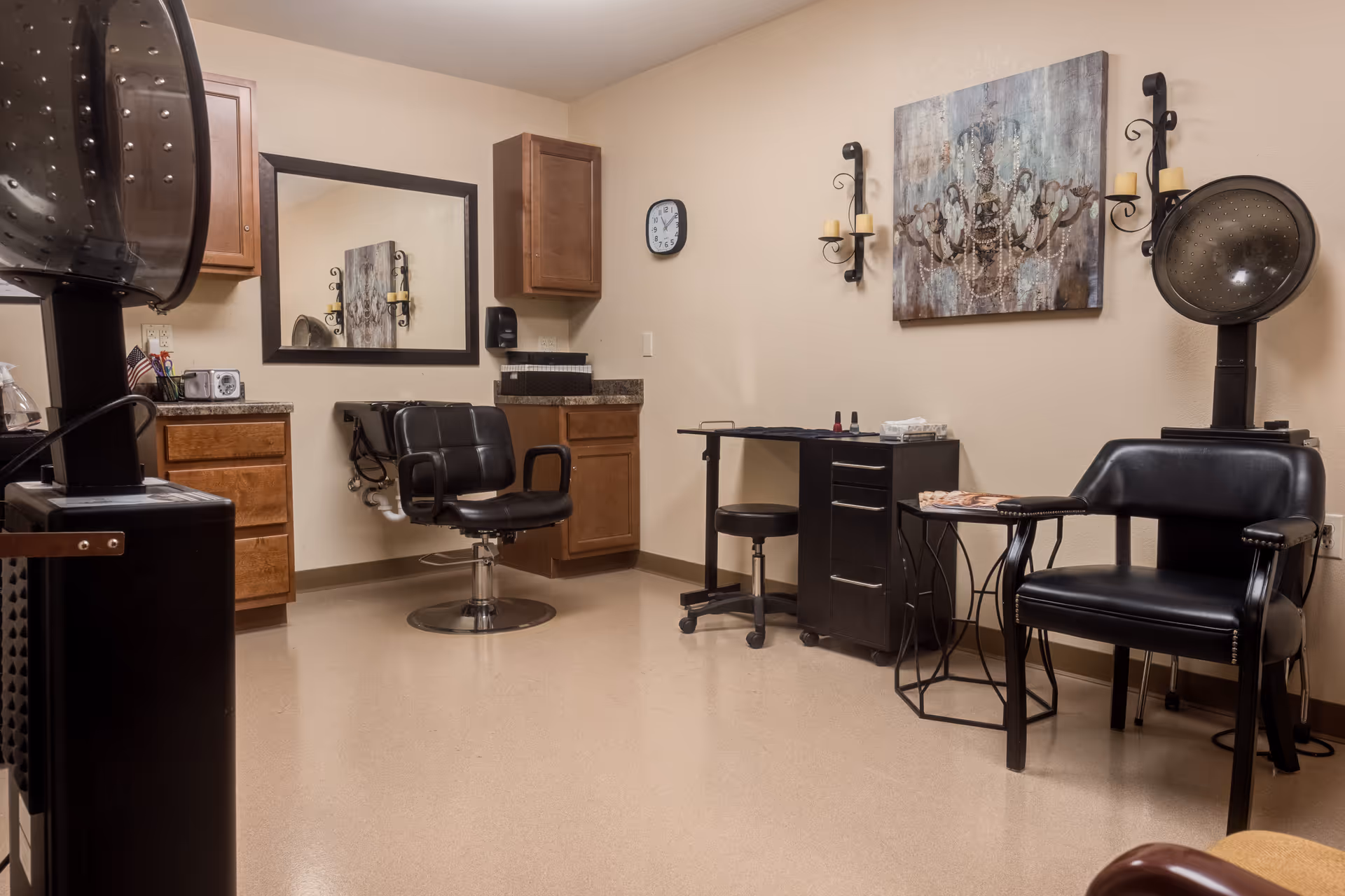 Interior view of a salon area in a senior living facility with two black salon chairs, hair drying machines, a manicure table with a stool, wooden cabinets, a wall clock, a large mirror, and decorative wall art with candle holders.