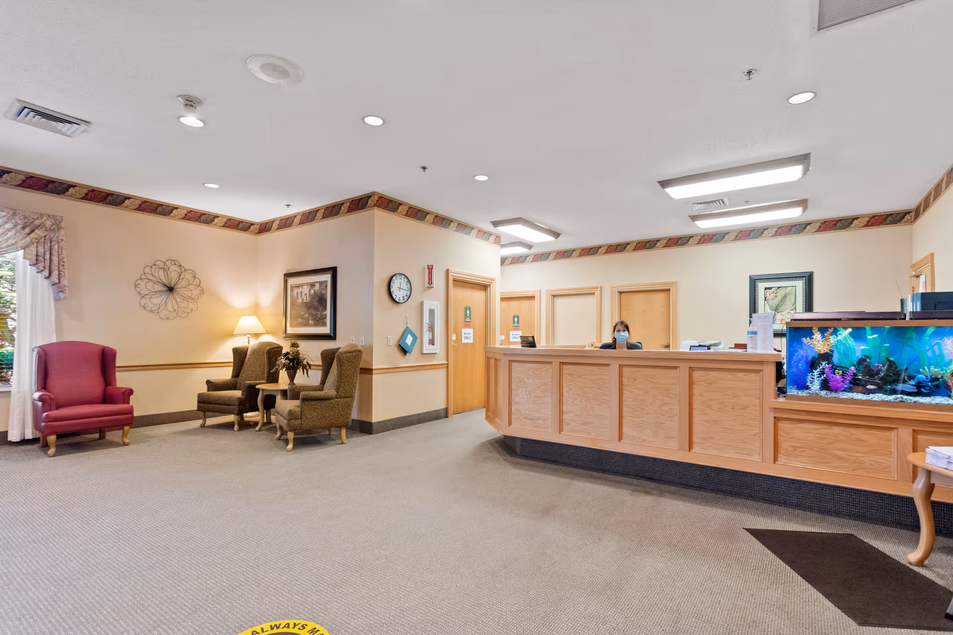 Reception area of Sycamore Run Nursing & Rehabilitation Center with a wooden front desk, an aquarium on the right side, a receptionist wearing a mask behind the desk, and a seating area with armchairs and a lamp on the left side. The room has beige walls with a decorative border near the ceiling and carpeted floor.