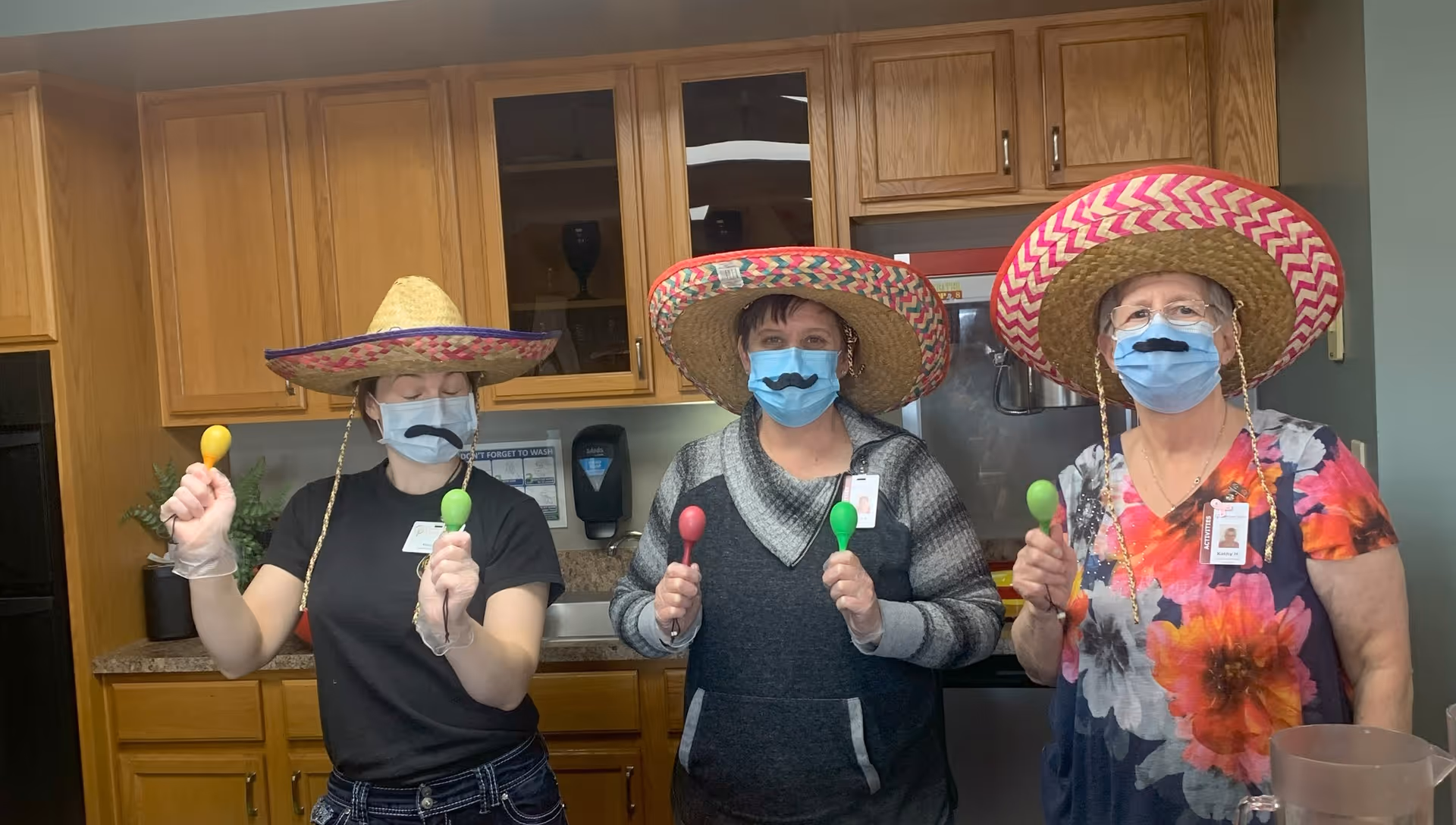 Three women wearing colorful sombreros and blue face masks with fake mustaches printed on them, each holding maracas. They are standing in a kitchen area with wooden cabinets and a countertop behind them.