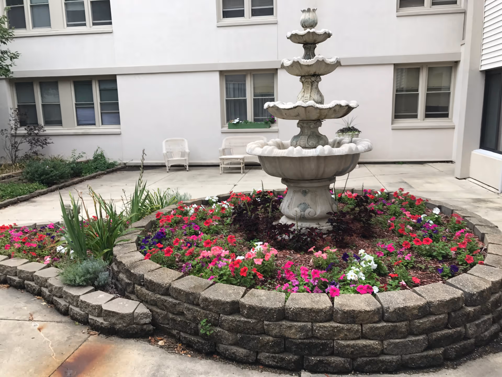 Outdoor courtyard area with a multi-tiered stone fountain surrounded by a circular flower bed filled with colorful flowers. In the background, there are white wicker chairs and a bench against a white building with several windows.
