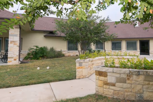 Outdoor view of a landscaped garden area with a curved stone planter filled with green plants, a tree, and a building with beige siding and multiple windows in the background.
