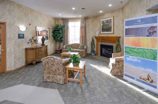 A cozy senior living facility common room with upholstered chairs and sofas around coffee tables, a fireplace, plants, and a wellness banner.